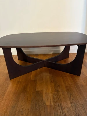 Mörkbrunt soffbord i trä - Säljer ett stilrent soffbord i mörkbrunt trä med rektangulär, rundad bordsskiva och unikt korsformat underrede. Bordet har en modern design som passar perfekt till vardagsrummet och ger en cool touch till inredningen.