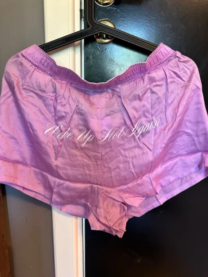 Lila saten pyjamasshorts från PINK - Säljer ett par lila pyjamasshorts från PINK med texten 'Wake Up Hot Again' på baksidan. Jag säljer den eftersom den är för stor för mig.