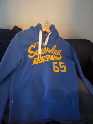 Blå Superdry hoodie med tryck - Snygg blå hoodie från Superdry med gul text 'Team Superdry Comets 65' på bröstet. Klassisk känguruficka framtill, vita dragsnören i huvan och mjukt material. Perfekt för en avslappnad och sportig stil.