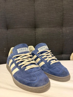 Blå Adidas Handball Spezial sneakers - Snygga Adidas Handball Spezial sneakers i blå mocka med vita detaljer och klassiska tre ränder på sidan. Skorna har vita snören och grå sula. Perfekta för dig som gillar retrostil och streetwear.