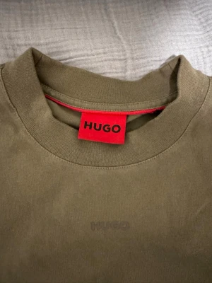 Olivgrön t-shirt från HUGO - Snygg olivgrön t-shirt från HUGO med diskret logga på bröstet och klassisk rund hals. T-shirten har korta ärmar och är tillverkad i mjuk bomull som känns skön mot huden. Perfekt för en clean och stilren look.