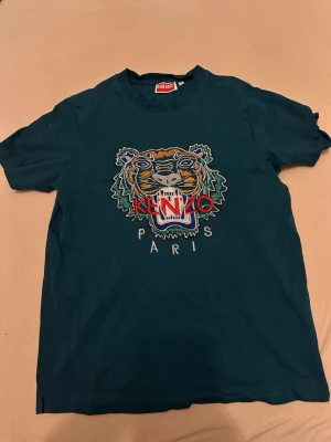 Blå Kenzo t-shirt med tigerbrodyr - Säljer en blå t-shirt från Kenzo med ikonisk tigerbrodyr och texten 'KENZO PARIS' framtill. T-shirten har korta ärmar och är gjord i mjuk bomull. Perfekt för dig som vill sticka ut med en snygg och trendig look.