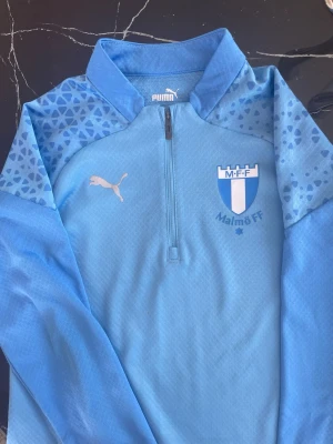 Ljusblå Malmö FF träningströja Puma - Snygg ljusblå träningströja från Puma med Malmö FF-logga på bröstet. Tröjan har halv dragkedja, ståkrage och mönstrade axelpartier i blått. Perfekt för fotbollsträning eller supporterliv. Utan skador 