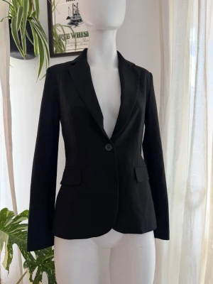 ZARA Svart klassisk kavaj med fickor - Stilren svart kavaj med en knapp framtill och två fickor med lock. Kavajen har smal passform, klassisk krage och långa ärmar. Perfekt för en clean och snygg look. Materialet ser ut att vara en blandning av bomull och polyester.