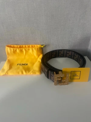 Fendi brunt bälte med FF-logga - Snyggt brunt bälte från Fendi med klassiskt FF-mönster i svart och brunt. Bältet har ett stort, guldigt Fendi-spänne med struktur. Perfekt accessoar för att lyfta din outfit och visa din stil.