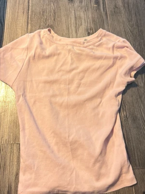 Ljusrosa Hello Kitty t-shirt - Söt ljusrosa t-shirt med korta ärmar och ett glittrigt Hello Kitty-motiv framtill i små stenar. T-shirten har en enkel, klassisk passform och är gjord i mjuk bomull som känns skön mot huden. Perfekt för dig som gillar gulliga detaljer och vill sticka ut lite extra. Tyvärr har ett par få stenar åkt ut men inget som syns