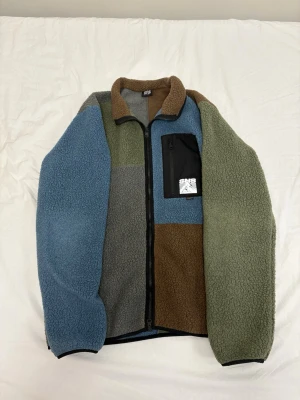 Fleece från SNS - Säljer en färgglad fleecejacka med patchwork-design i blått, grönt, brunt och grått. Jackan har hel dragkedja, hög krage och en svart bröstficka med dragkedja och SNS-logga. Materialet är mjukt teddyfleece som håller dig varm och bekväm.