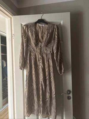 Adoore zebraklänning - Snygg långklänning med zebramönster i beige och svart. Klänningen har v-ringning, långa ärmar och knappar framtill. Använd fåtal gånger därav söndrig knapp.