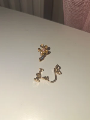 Guldiga örhängen med blomdetalj - Säljer ett par guldiga clipsörhängen med en söt blomformad design. Blomman har en liten glittrande sten i mitten som ger extra bling. Perfekta för dig som vill ha något unikt och elegant till din stil. Aldrig använt, och är som nytt. Erbjuder dessa tills den 19 Juli, vi kan diskutera pris, om det behövs. Tack.