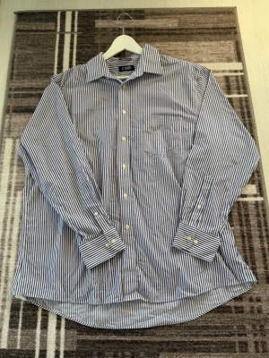 Randig blåvit skjorta från Chaps - Klassisk långärmad skjorta från Chaps i blått och vitt med smala ränder. Skjortan har knappar framtill, bröstficka och normal passform. Perfekt till jeans eller chinos för en clean look.