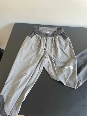 Grå mjukisbyxor The North Face XS - Grå mjukisbyxor från The North Face med elastisk midja och meshfoder. Byxorna har logga på vänster ben, ribbade muddar vid bensluten och är tillverkade i ett lätt och bekvämt material. Perfekta för träning eller chill. 