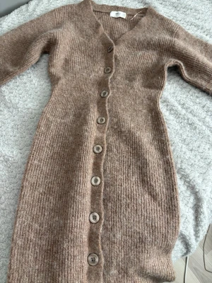 Beige stickad klänning med knappar - Mysig beige stickad klänning med v-ringning och långa ärmar. Klänningen har ribbad struktur och knappar hela vägen framtill, vilket ger en snygg och avslappnad look.