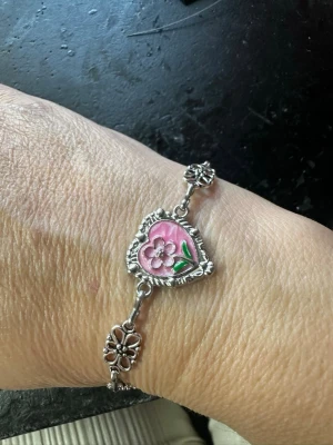 Silverfärgat armband med hjärta och blomma - Unikt armband i silverfärgad metall med ett hjärtformat hänge i mitten. Hänget har en rosa bakgrund och en upphöjd blomma med gröna blad. Länkarna består av dekorativa blommor som ger en vintagekänsla. Perfekt för dig som gillar romantiska detaljer och färg.