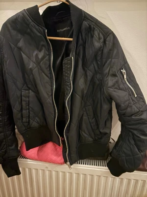 Svart quiltad bomberjacka Rut&Circle - Svart bomberjacka från Rut&Circle med quiltat mönster, ribbade muddar och silverfärgad dragkedja. Jackan har fickor på sidorna och en ärmficka med dragkedja. Perfekt för en cool och avslappnad stil.