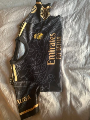 Svart Real Madrid matchtröja - Snygg svart matchtröja från Real Madrid med gulddetaljer och drakmönster. Tröjan har korta ärmar, rund hals och tryckta loggor som 'Emirates Fly Better' och 'La Liga'. Perfekt för fotbollsträning eller att bära till vardags.