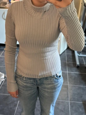 Ljusgrå ribbad polotröja - Säljer en ljusgrå ribbstickad polotröja med lång ärm och figurnära passform. Tröjan har hög krage och är tillverkad i ett stretchigt material som sitter snyggt på kroppen. Perfekt att styla med jeans eller kjol. I storlek S.