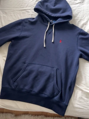 Ralph lauren hoodie - Snygg mörkblå hoodie med vit dragsko och klassisk känguruficka framtill. På bröstet finns en liten röd broderad logga. Hoodien har en avslappnad passform och är tillverkad i mjukt bomullsmaterial. Perfekt för en chill och stilren look.