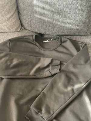 Svart basic sweatshirt - En enkel svart sweatshirt med rund halsringning och ribbade muddar vid ärmar och nederkant. Tröjan har en clean look utan tryck eller logga och är gjord i ett mjukt material som känns skönt mot huden. Perfekt för en avslappnad stil.