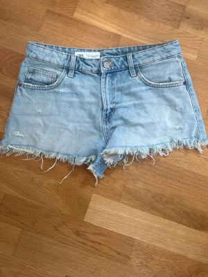 Ljusblå lågmidjade jeansshorts med fransar - Säljer ett par lågmidjadeljusblå jeansshorts med råa, fransiga kanter och slitna detaljer. Klassisk femficksmodell med knapp och dragkedja framtill. Perfekta för sommaren och ger en avslappnad vibe. 