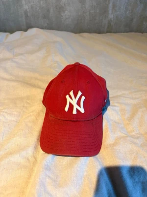 Röd New York Yankees keps - Snygg röd keps med New York Yankees-logga i vitt framtill. Klassisk böjd skärm och broderad New Era-logga på sidan. Justerbar passform och tillverkad i bomullstyg. Perfekt för att lyfta din streetwear-look.