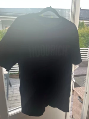 Svart HOODRICH t-shirt - Säljer en svart t-shirt från HOODRICH med tryck både fram och bak. T-shirten har klassisk passform och korta ärmar. Perfekt för dig som gillar streetwear och vill ha en enkel men snygg look.