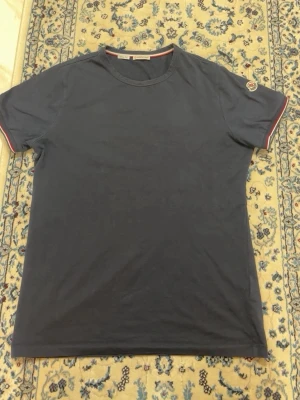 Mörkblå Moncler t-shirt med logga - Snygg mörkblå t-shirt från Moncler med korta ärmar och klassisk rund hals. Ärmsluten har rödvita ränder och Moncler-logga på vänster ärm. Tillverkad i mjuk bomull för en skön känsla.