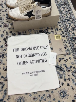 Golden Goose vita sneakers med svart stjärna - Snygga vita sneakers från Golden Goose med svart stjärndetalj på sidan och beige snören. Skorna har en klassisk låg siluett, perforerade detaljer på tån och beige sula för en vintage look. Tillverkade i läder med svarta detaljer på häl och plös.
