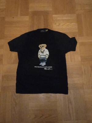 Svart Polo Bear t-shirt från Ralph Lauren - Svart t-shirt från Ralph Lauren med ikoniska Polo Bear-trycket på bröstet. Klassisk rund hals och korta ärmar. Perfekt för dig som gillar streetwear och vill ha en tidlös look med en twist.
