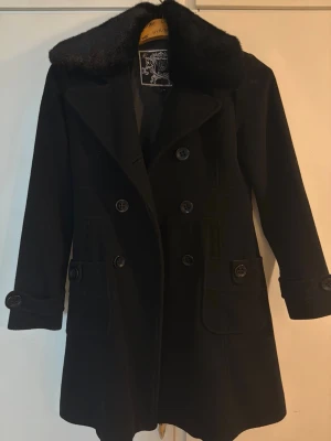 Svart kappa med fuskpäls -  svart kappa med dubbelknäppning och med päls. Kappan har fickor med dekorativa knappar och markerad midja för en snygg siluett. Perfekt för kyliga dagar och ger en klassisk look.