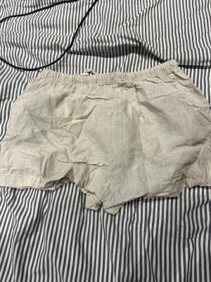 Beige linneshorts från H&M - Säljer ett par ljusa, beige shorts från H&M i luftig linnemix. De har elastisk midja och en avslappnad passform, perfekta för varma dagar. Enkla att matcha med allt och superbekväma att ha på sig.