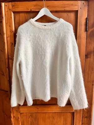 Vit fluffig stickad mohair tröja - Mysig vit stickad tröja med fluffig yta och rund halsringning. Tröjan har en avslappnad passform och långa ärmar, perfekt för kyliga dagar. Materialet ser ut att vara en mjuk blandning med mohair eller liknande för extra komfort.