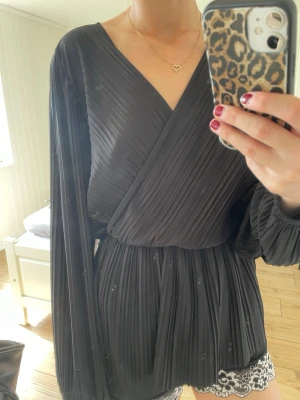  Svart omlottblus - Snygg svart omlottblus med plisserat tyg och långa ärmar. Blusen har v-ringning och resår i midjan. Blusen är från H&M i storlek xs.