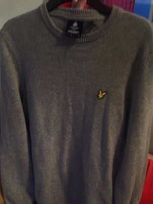 Grå stickad tröja från Lyle & Scott - Snygg grå stickad tröja från Lyle & Scott med rund halsringning och den klassiska gula logon broderad på bröstet. Tröjan har långa ärmar och ribbade muddar vid ärmslut och nederkant. Perfekt för lager på lager och enkel att matcha.
