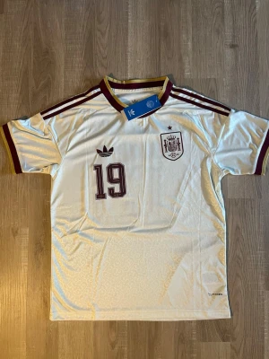 Vit Spanien fotbollströja  - Säljer en vit Spanien fotbollströja med vinröda detaljer, nummer 19 och Lamine Yamal på ryggen. Tröjan har korta ärmar, rund hals och klassiska ränder på axlarna. HELT NY!