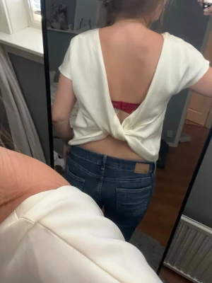 Vit omlottopp med öppen rygg - Säljer en vit topp i satinliknande material med korta ärmar och snygg omlottdetalj bak. Toppen har öppen rygg och en croppad passform som ger en trendig look. Perfekt för dig som vill sticka ut lite extra! Liten ”reva” på framsidan av lappen, se bilder i annonsen.