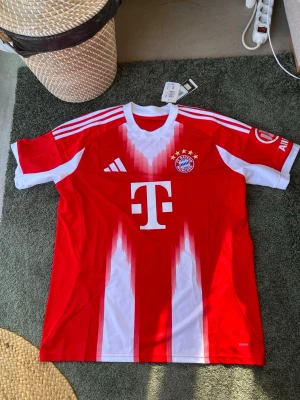 FC Bayern München matchtröja röd - Säljer en officiell FC Bayern München matchtröja i rött och vitt med spelarnamn Olise och nummer 17 på ryggen. Tröjan har klubbmärke, Adidas-logga och T-Mobile sponsortryck på framsidan. Tillverkad i lätt och ventilerande material, perfekt för fotbollsträning. Tröjan är helt ny och aldrig burits.