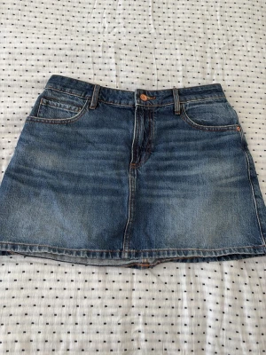 Blå jeanskjol med fickor - Säljer en klassisk blå jeanskjol i kort modell med fem fickor och bälteshällor. Kjolen har en rak passform och stängs med knapp och dragkedja framtill. Perfekt till vardags för en avslappnad och trendig look.
