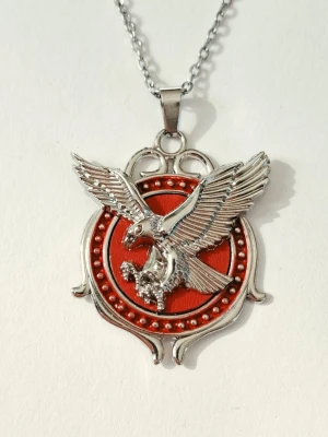 Halsband med silverfärgad örn - Snyggt halsband med ett stort silverfärgat örnhänge mot en röd bakgrund. Hänget har detaljerade vingar och en dekorativ ram med små kulor runt den röda emalj cirkeln. Kedjan är i metall. Materialet rostfritt stål 