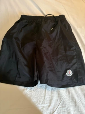 Svarta Moncler shorts med snörning - Säljer ett par svarta shorts från Moncler med elastisk midja och snörning. Shortsen har en Moncler-logga på vänster ben och praktiska fickor, inklusive en bakficka med knapp. Tillverkade i ett lätt och glansigt syntetmaterial, perfekta för sommaren.