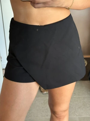 Svart omlott shorts - Snygg svart skort med omlottdesign framtill. Skorten har en hög midja och korta ben, vilket ger en modern och stilren look. Tillverkad i ett mjukt och stretchigt material som sitter bekvämt på kroppen. Perfekt för dig som vill ha både kjol- och shortslook.