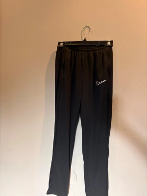 Svarta träningsbyxor från Nike - Svarta mjukisbyxor från Nike med vit broderad Swoosh-logga på vänster ben. Byxorna har resår i midjan och raka ben. Perfekta för en avslappnad stil och tillverkade i mjukt material för extra komfort.