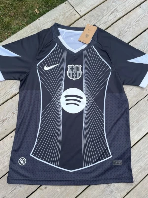 Svart FC Barcelona matchtröja Nike - Snygg svart FC Barcelona matchtröja från Nike med vita grafiska linjer och klubbmärke på bröstet. Tröjan har korta ärmar, rund hals och vit Spotify-logga framtill. Tillverkad i lätt och ventilerande material, perfekt för fotboll, träning eller i vardagen 