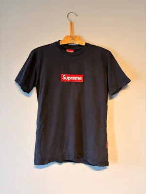Svart Supreme t-shirt med box logo - Supreme t-shirt i svart med den klassiska röda box logon broderad på bröstet. T-shirten har rund hals och korta ärmar. Perfekt för dig som gillar streetwear och vill ha en ikonisk look.