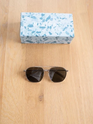 Silvriga aviator pilot solglasögon Nividas - Snygga solglasögon i klassisk aviator-modell med tunna silvriga metallbågar och mörka, rektangulära linser. Perfekta för att höja din stil på sommaren. Kommer med ett stilrent fodral i blått och vitt mönster.