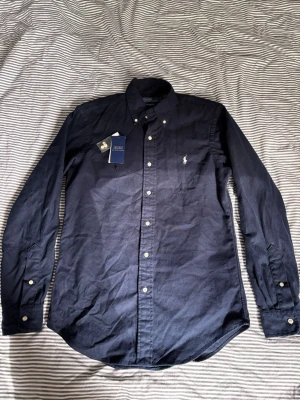 Mörkblå skjorta från Polo Ralph Lauren - Klassisk mörkblå skjorta från Polo Ralph Lauren med button-down krage och vit broderad logga på bröstet. Skjortan har långa ärmar, vita knappar och är tillverkad i mjuk bomull. Perfekt för en stilren look.