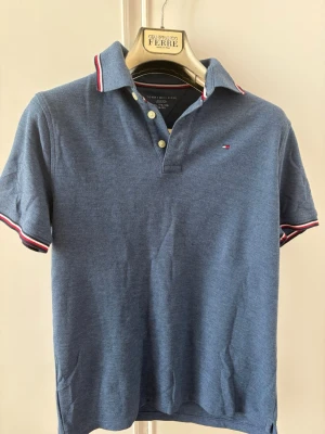 Blå pikétröja från Tommy Hilfiger - Snygg blå pikétröja från Tommy Hilfiger med klassisk krage och knappar framtill. Tröjan har korta ärmar och detaljer i rött, vitt och blått på krage och ärmslut. Liten logga på bröstet. Perfekt till en avslappnad stil.  Storlek (16 - 18 ) Xl junior helt ny! Oanvänd 