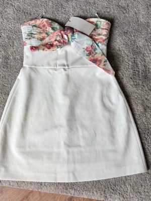 Vit kort klänning med blommig överdel  - Snygg axelbandslös kort klänning i vitt med en draperad, blommig överdel i pastellfärger och rosettdetalj framtill. Klänningen har en feminin siluett och är perfekt för sommarens alla tillfällen. Dragkedja baktill för enkel påklädning.helt ny och populär