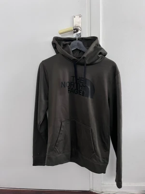 Mörkgrön hoodie från The North Face - Snygg mörkgrön hoodie från The North Face med stor logga på bröstet. Hoodien har huva med svarta dragsnören och en klassisk känguruficka framtill. Tillverkad i mjukt material som känns skönt mot huden. Perfekt för en avslappnad streetwear-look.