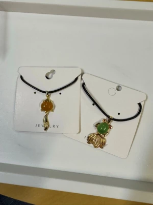 Två halsband med olika färger jade - Säljer två halsband med svarta band och hängsmycken i form av blommor. Ena halsbandet har en orange jade med guldfärgad blomdetalj, det andra har en grön jade med guldfärgad blomma. Perfekta accessoarer för att lyfta din stil.