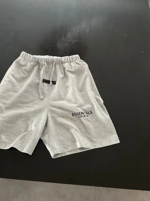 Grå shorts Essentials Fear of God - Snygga ljusgrå shorts från Essentials Fear of God med elastisk midja och snörning. Shortsen har en relaxed passform och diskret logga på vänster ben. Perfekta för chill dagar och enkel stil.